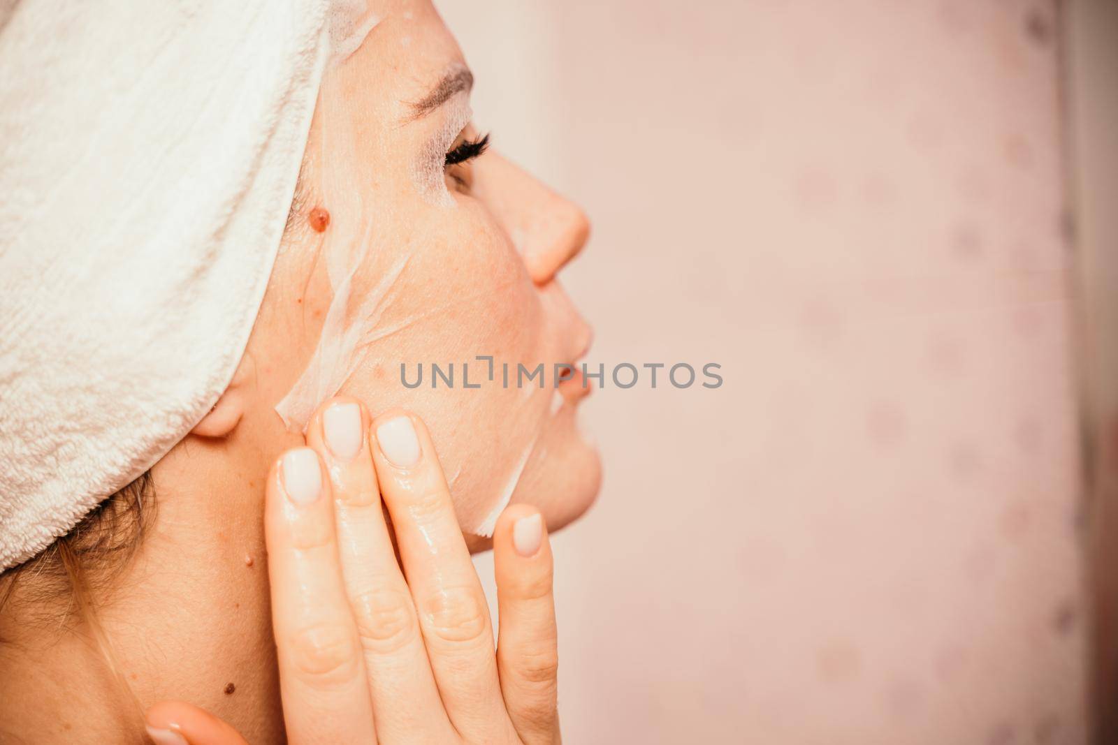 Young beautiful woman using a moisturizing facial mask after taking a bath. Pretty attractive girl in a towel on her head stands in front of a mirror in a home bathroom. Daily hygiene and skin care.