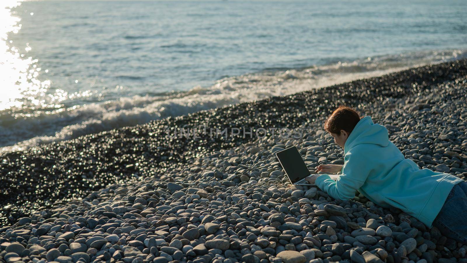 Caucasian woman lying on her stomach on a pebble beach and typing on a laptop. Remote work freelancer