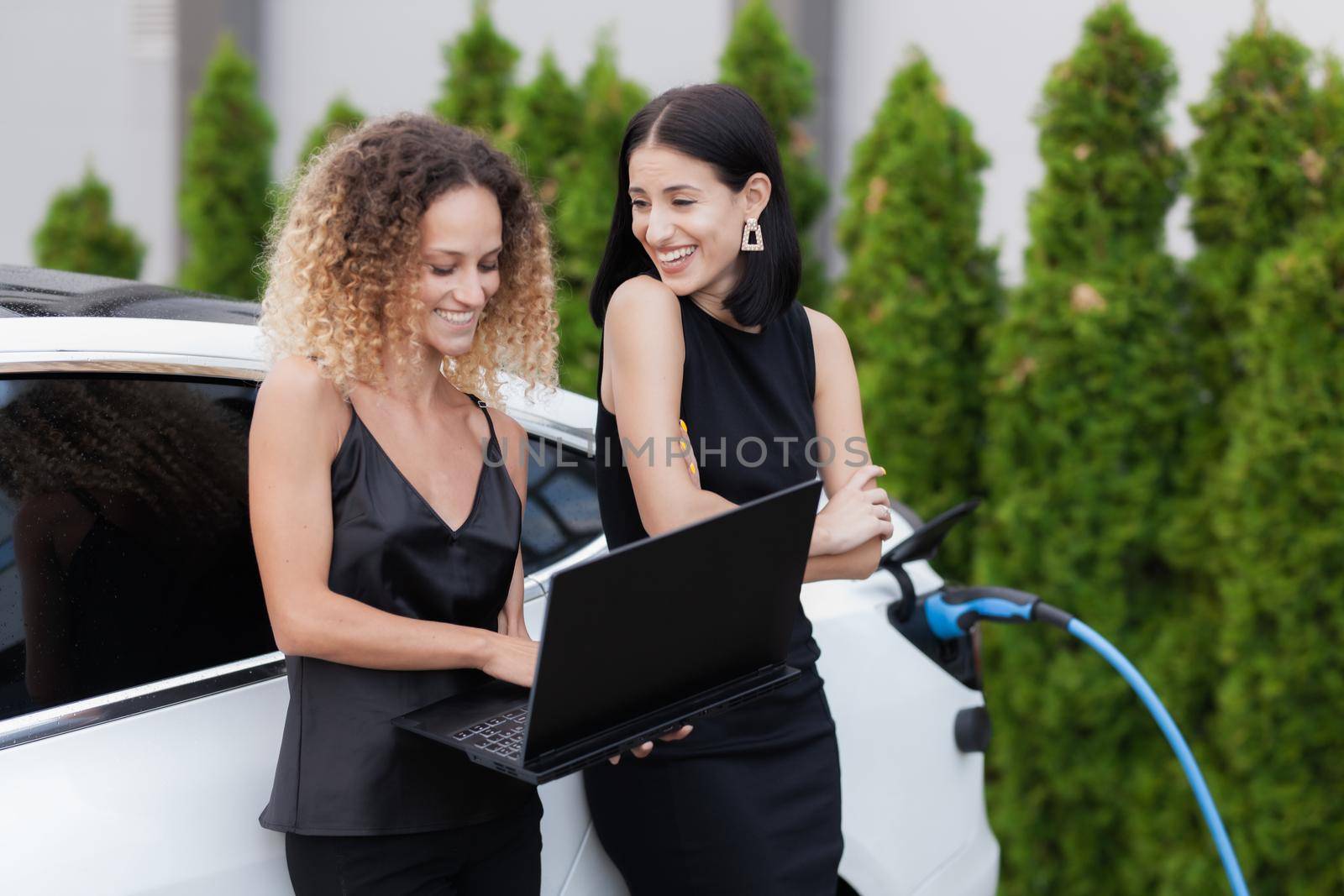 Two beautiful girls in black outfut smiling and posing. Businesswomen working on laptop while their electric car is charging in the background.