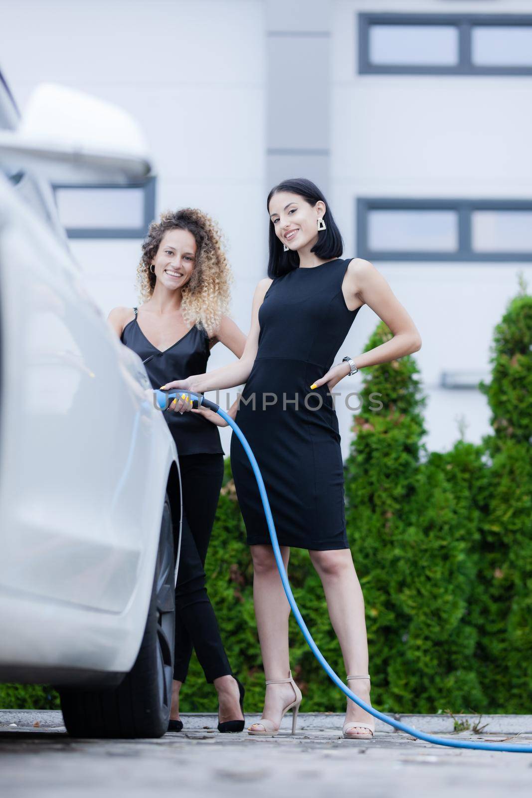 Two beautiful businesswomen in black outfut posing next to an electric car. Girls plugging blue cable in power charging socket.. by kokimk