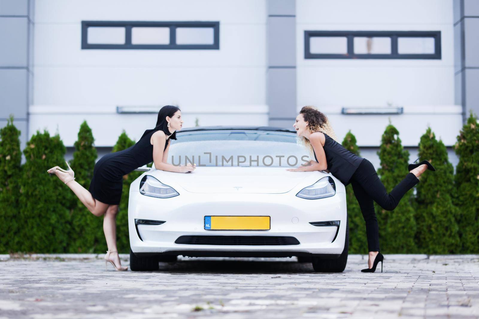 Two beautiful girls black outfut posing with modern white car. Urban wall background.. by kokimk
