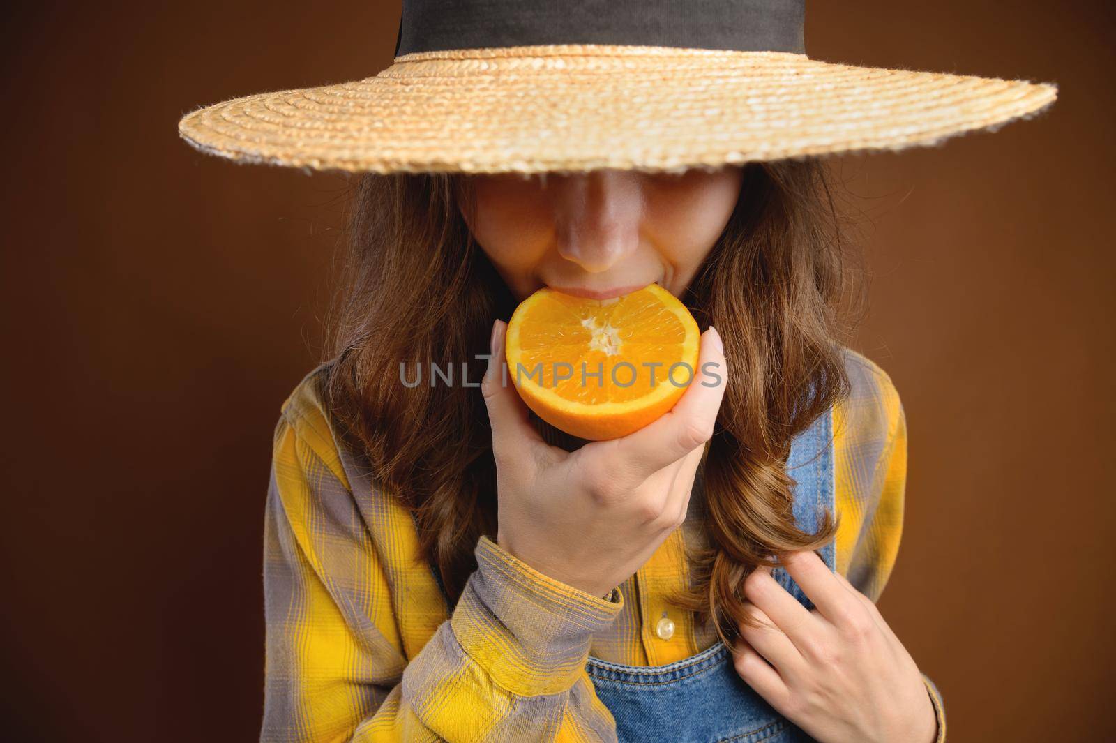 Studio portrait of an attractive caucasian young woman in a hat and denim overalls holds a cut orange fruit. in her hands and shows her tongue trying to taste it.