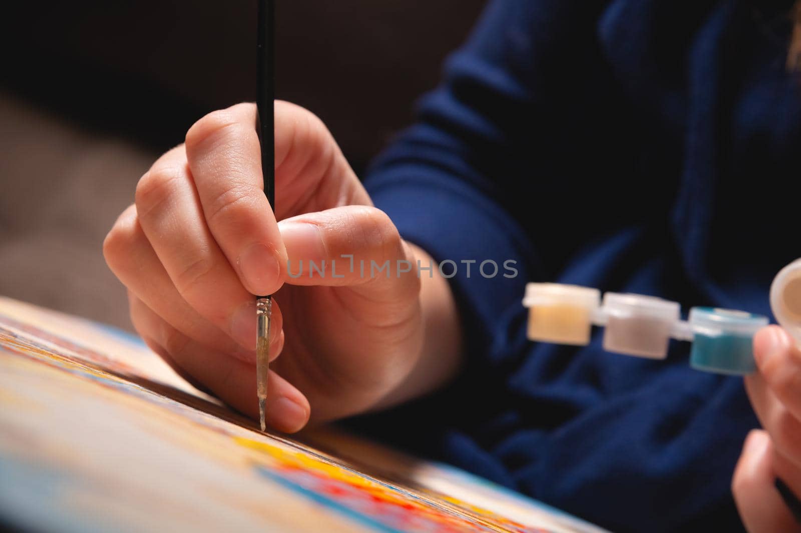 Close-up of a young female artist's hand with a thin brush painting a picture on canvas in a dark room. Shallow depth of field. high contrast.