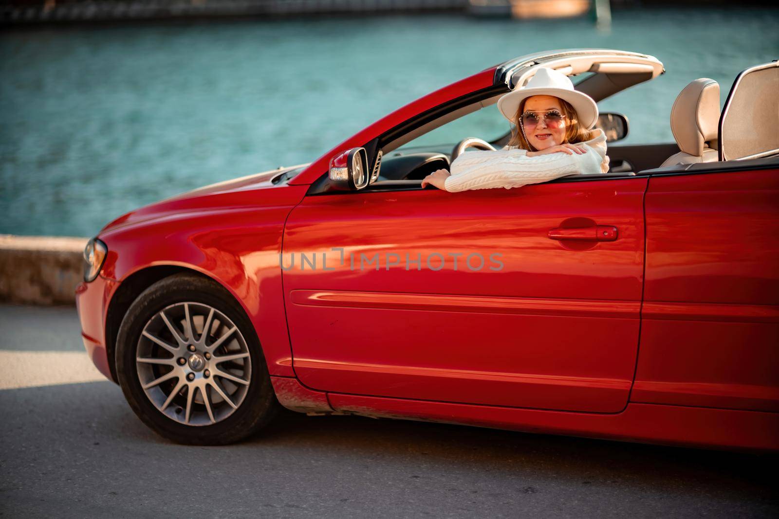 Outdoor summer portrait of stylish blonde woman driving red car convertible. Fashionable attractive woman with blond hair in a white hat in a red car. Sunny bright colors taken outdoors against the sea. by Matiunina