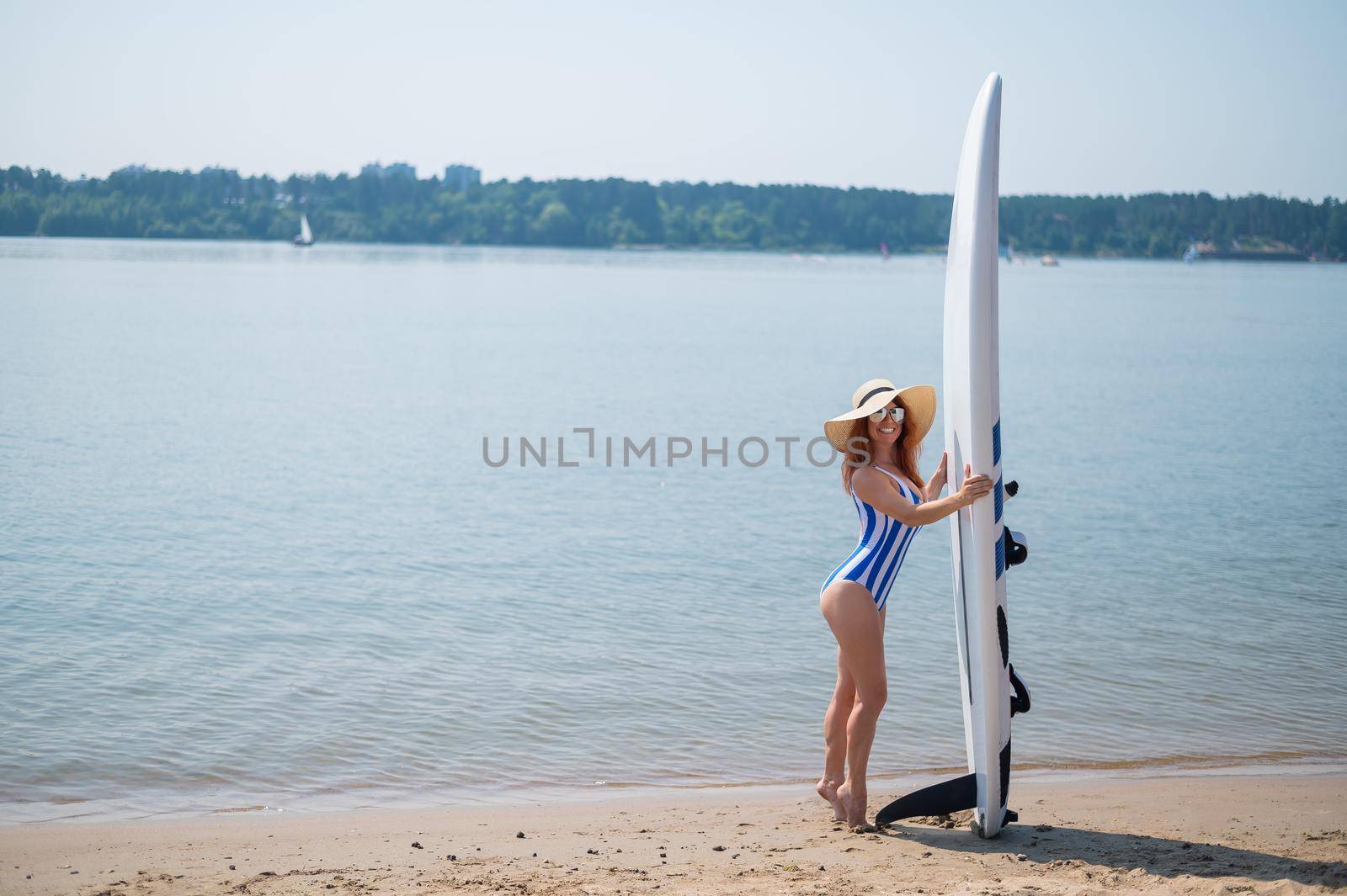 A beautiful red-haired woman in a straw hat, sunglasses and a striped one-piece swimsuit stands on the beach and holds a sup board. Happy smiling girl surfing on the lake. by mrwed54