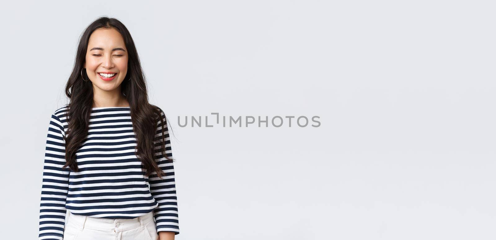 Lifestyle, people emotions and casual concept. Carefree, happy pretty asian woman in striped shirt close eyes and laughing sincere, having fun, funny moment, standing white background.