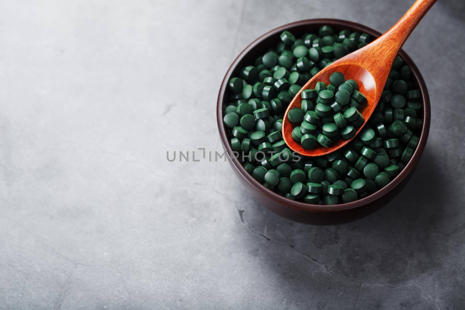 Organic spirulina tablets in a wooden cup with a wooden spoon on a dark background with free space top view