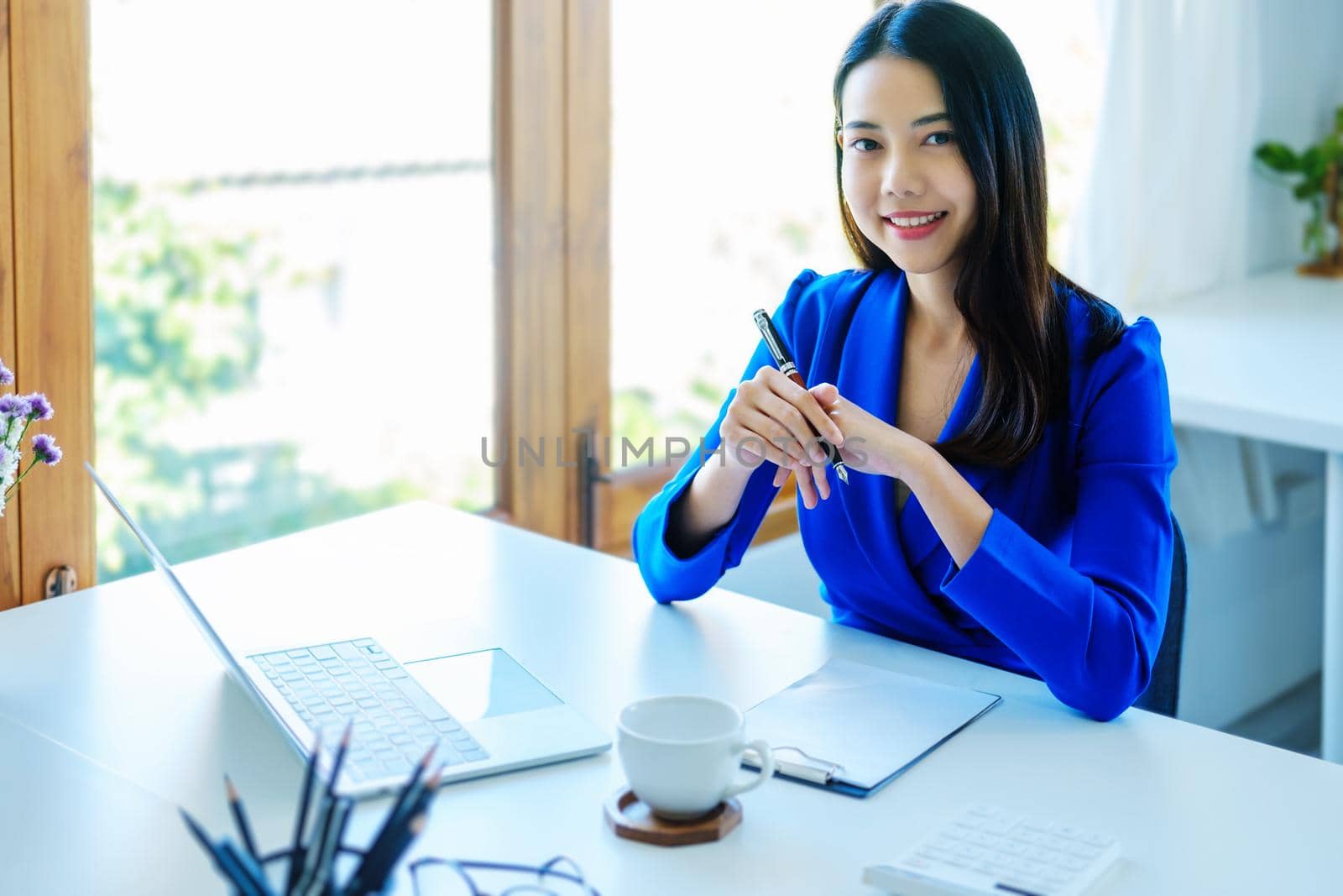 An Asian entrepreneur or businesswoman shows a smiling face while working with using computer on a wooden table. by Manastrong