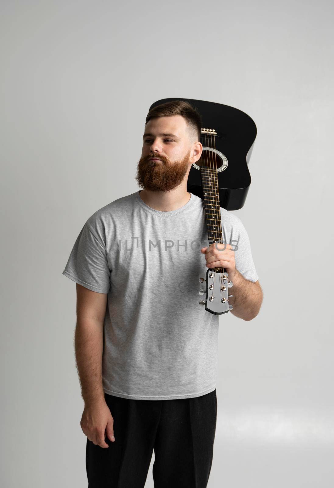 Handsome brunette bearded man musician standing and holding a acoustic guitar on a shoulder on a white background studio