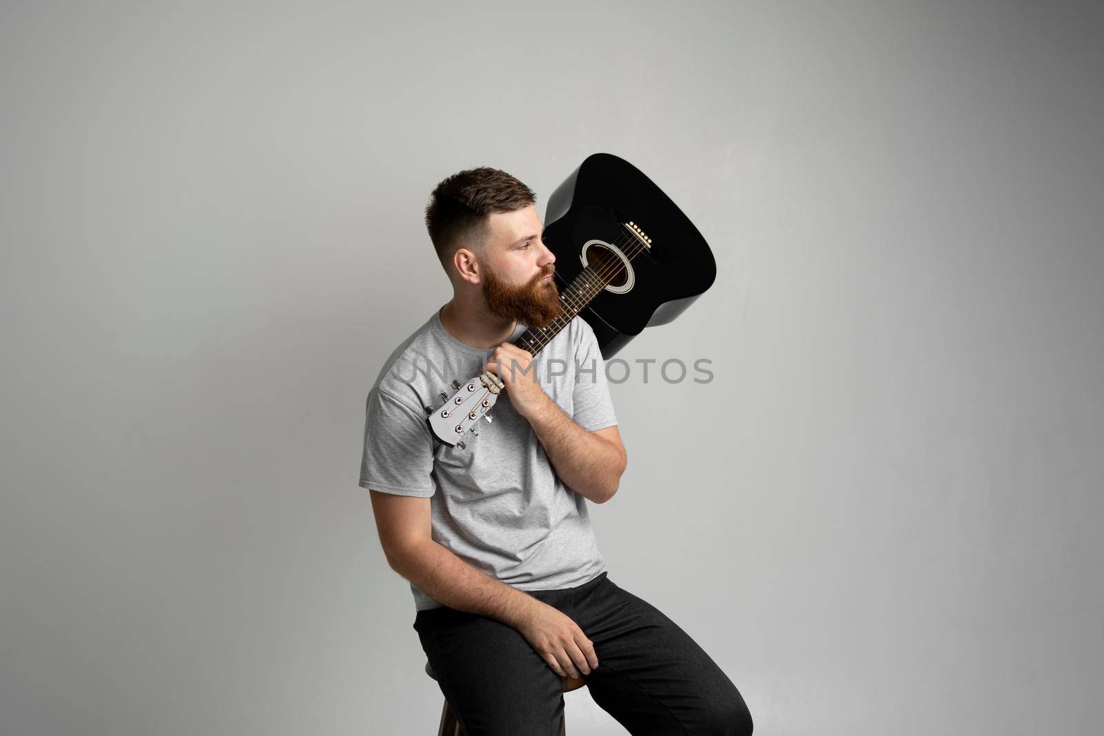 Handsome bearded brutal man musician in a grey t-shirt holding a acoustic guitar on a shoulder on a white background. by vovsht