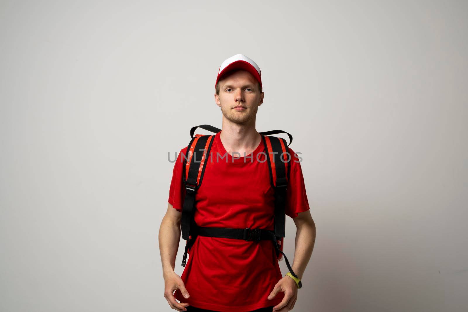 Young courier in a red uniform t-shirt and with red food thermo bag on a shoulder standing isolated on white background studio. Food delivery service