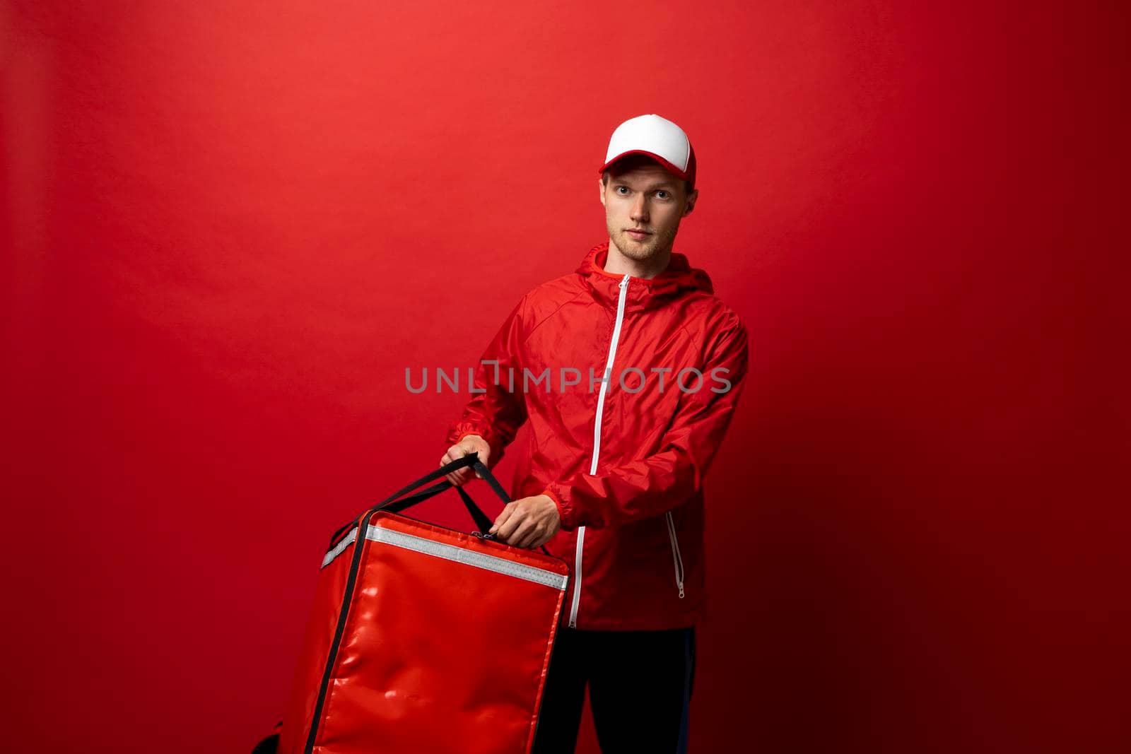 Food delivery service. Young courier in a red uniform and with a thermo bag on color red background, space for text