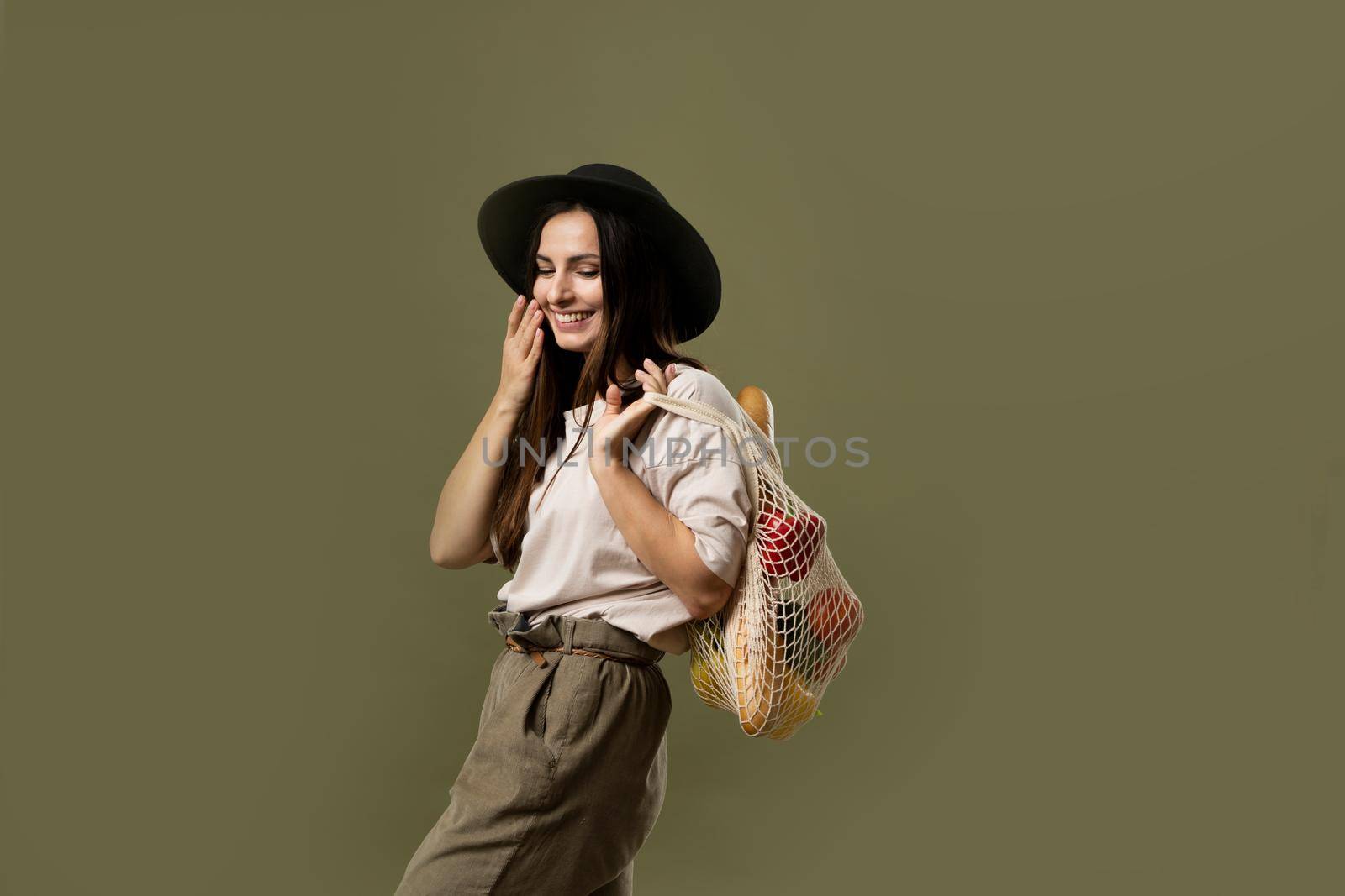 Zero waste concept. Young brunette woman in beige t-shirt and black hat holding mesh eco bag with organic fruits and vegetables. Using reusable crochet net bag for grocery shopping