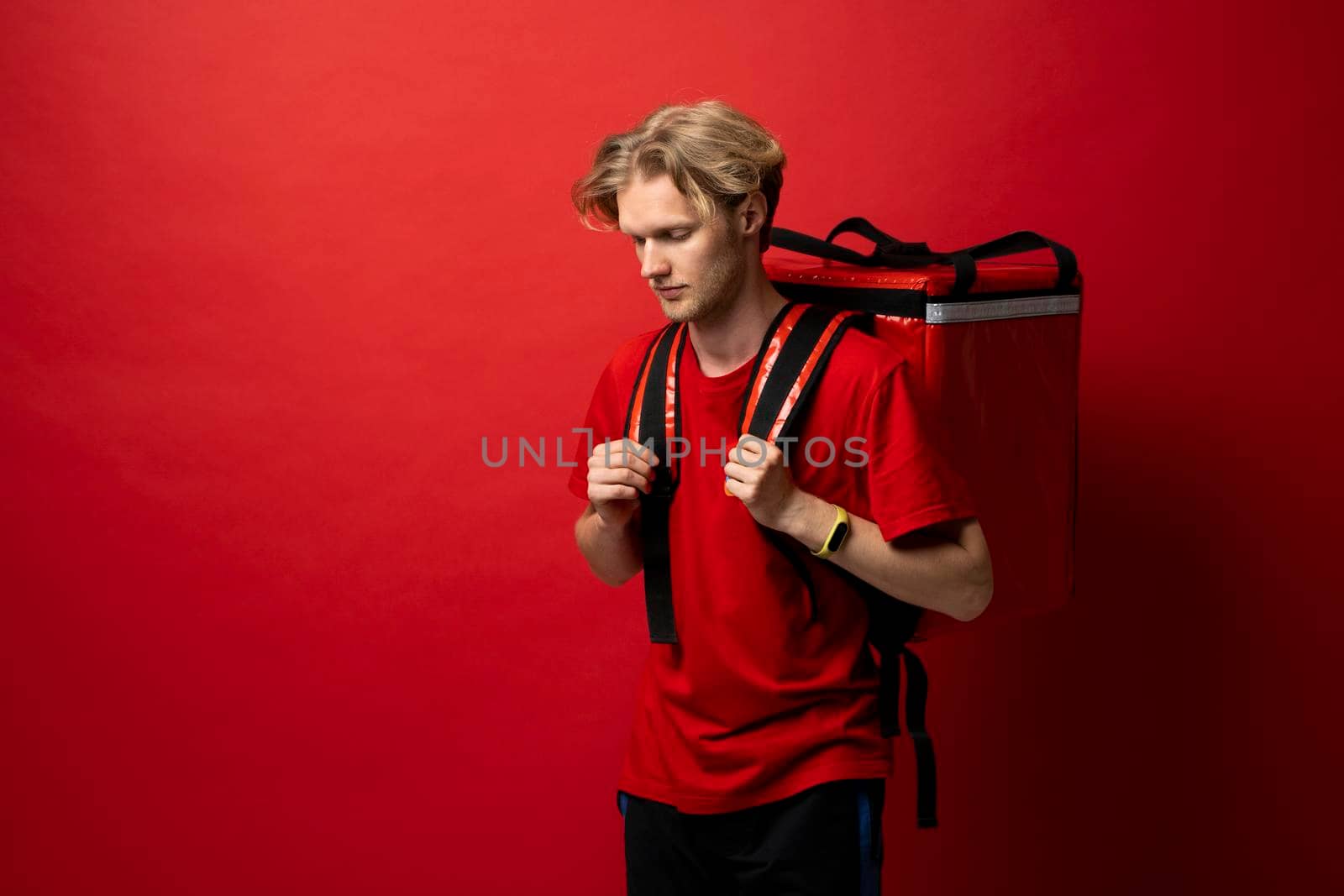 Young delivery employee man in red tshirt uniform with a thermal food bag backpack works as a courier in a food delivery service on a white background. by vovsht