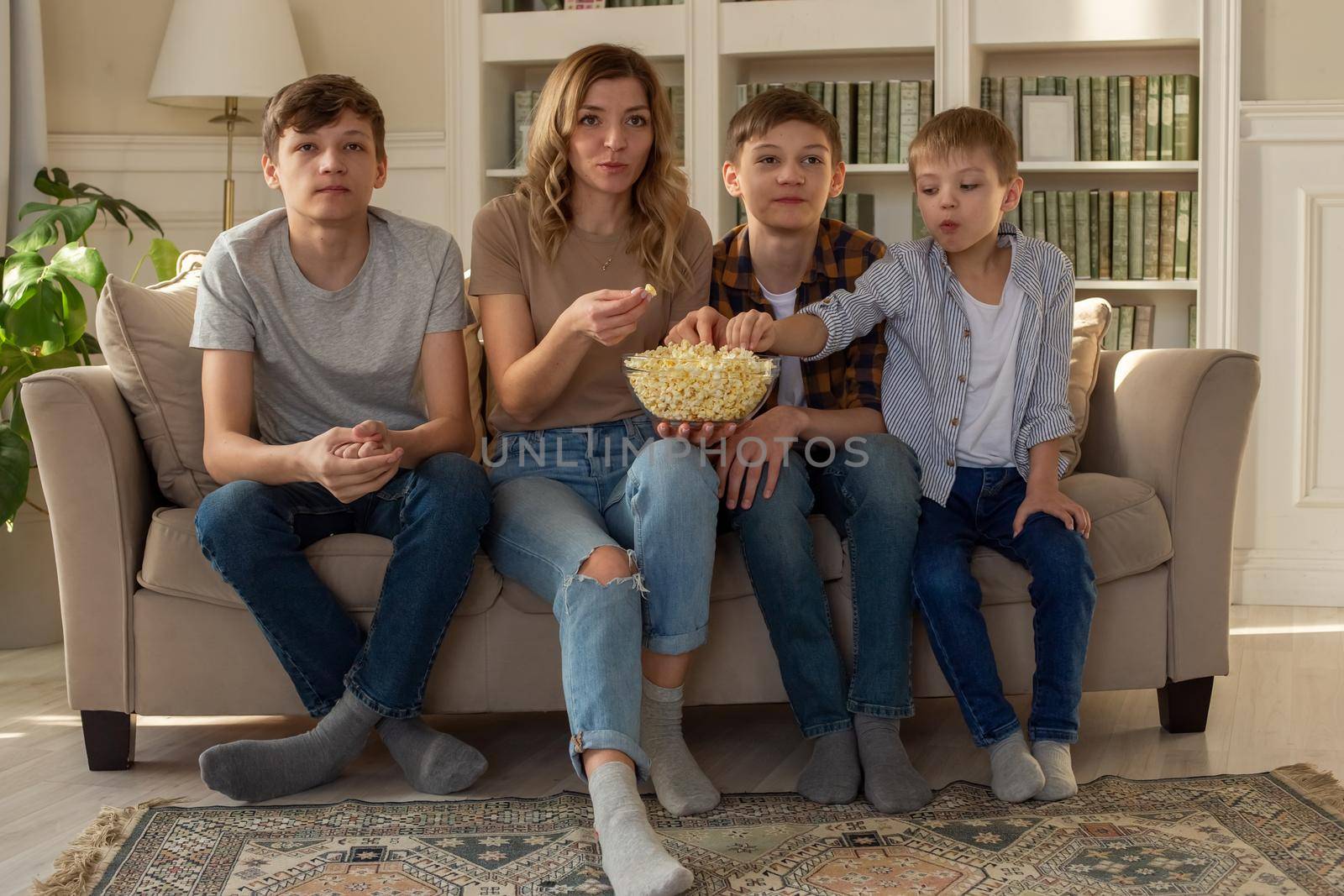 Happy family, a woman and three boys, are sitting on the sofa in the living room with a bowl of popcorn, watching TV by Zakharova