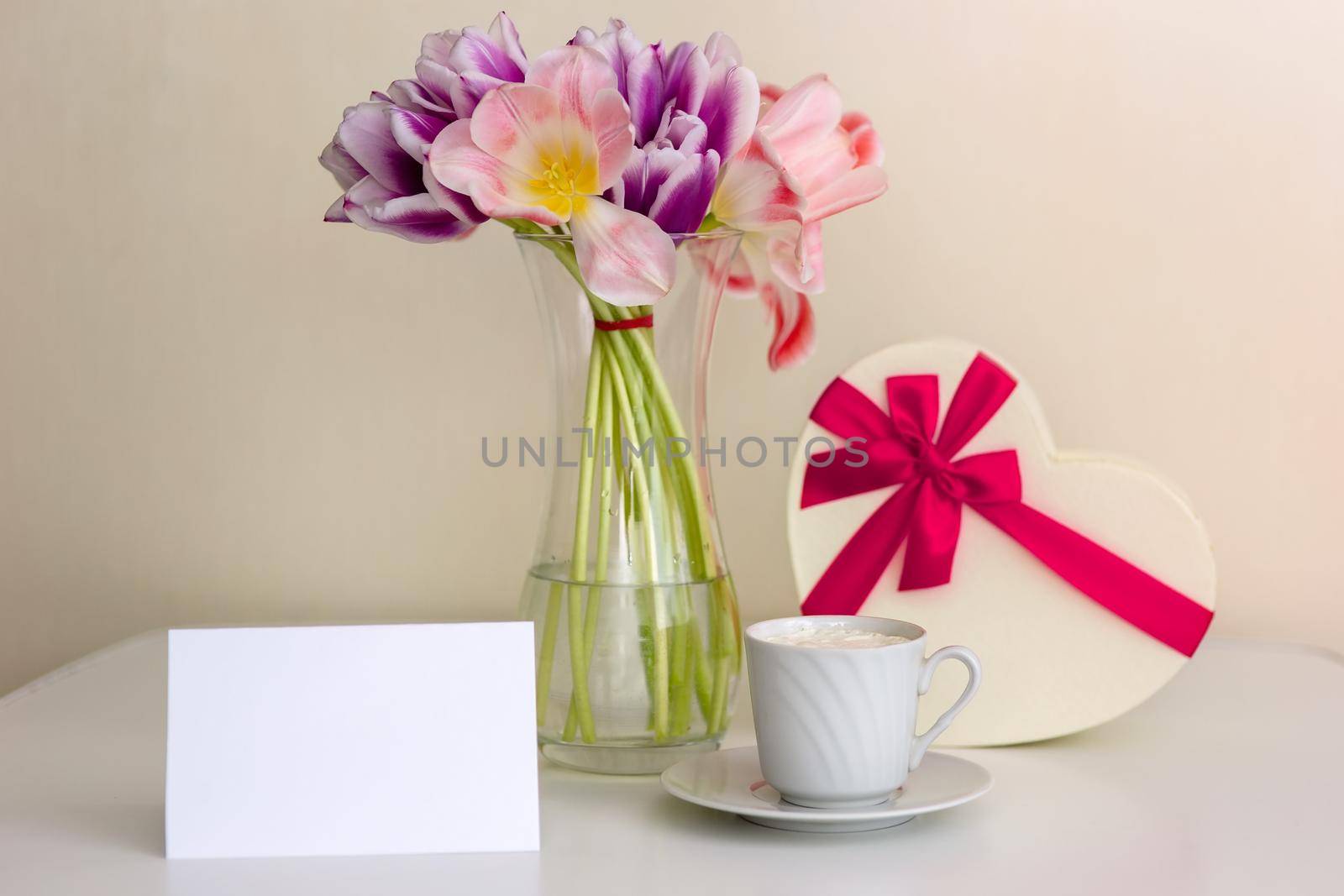 A bouquet of open pink and purple tulips, a gift box in the shape of a heart, with a pink bow, a white cup and a blank card with space for text on a white table in a bright room.