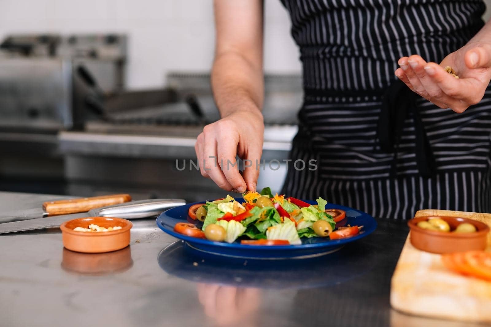 Detail of the hands of a young man, dressed in a black polo shirt and a dark striped apron, placing a salad on a blue plate in a professional kitchen, on a table full of food. The atmosphere is cold and clean, with the backdrop of professional aluminium tables and the fires of the restaurant kitchen.