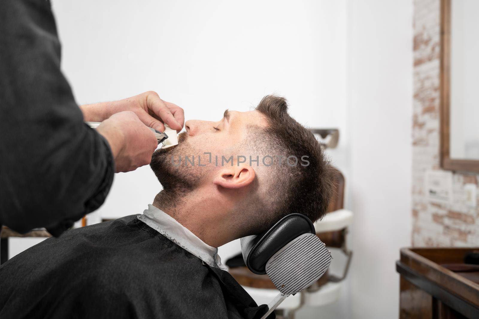 Young hipster Caucasian man during beard grooming in modern barber shop. Men's hair styling. Handsome man getting new hairstyle with electric trimmer. by HERRAEZ
