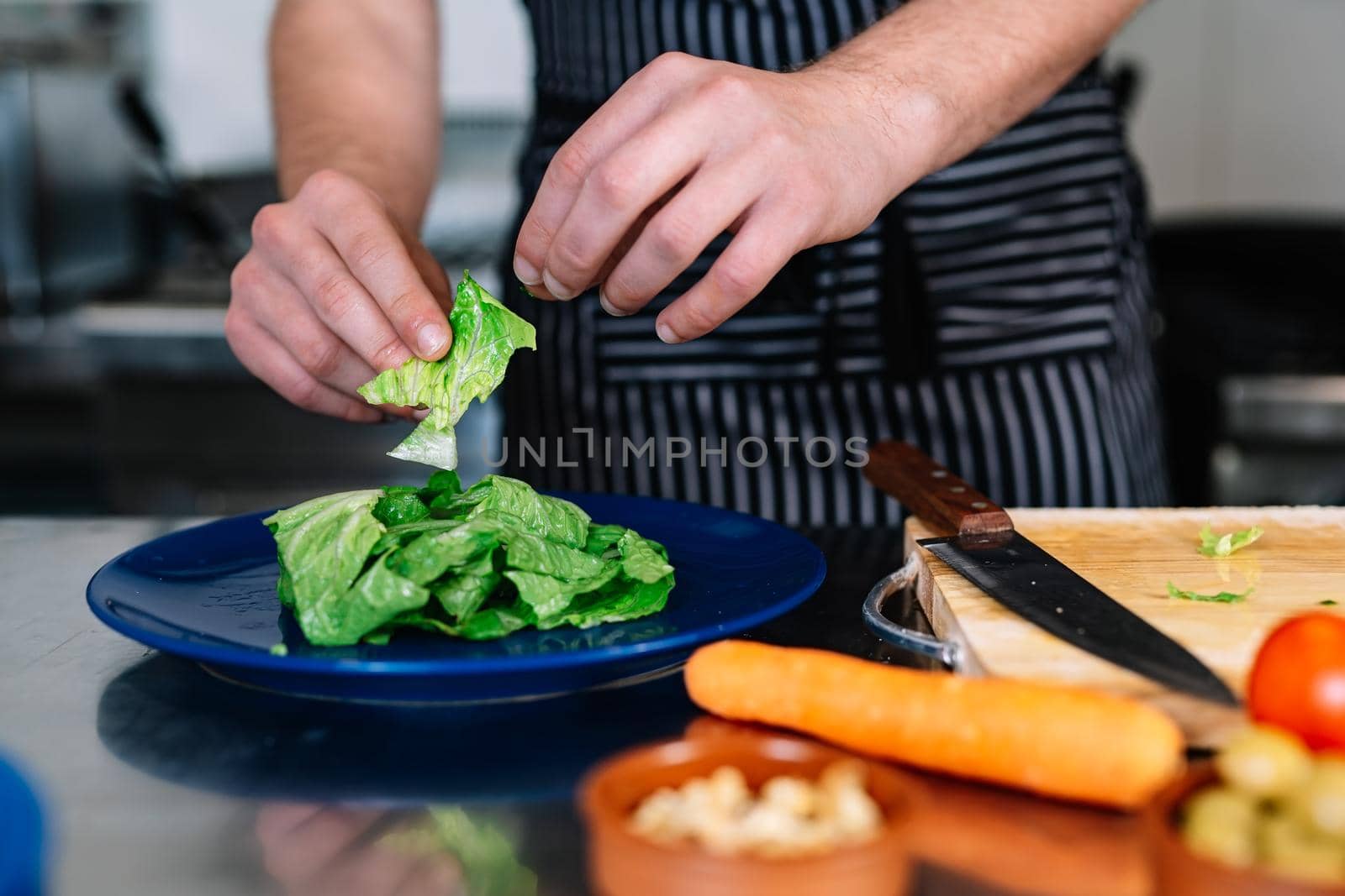 detail of the hands of a young man, dressed in a black polo shirt and a dark striped apron, preparing a salad dish in a professional kitchen, on a table full of food. Cold and clean environment, background of professional aluminium tables and cooking fires of a restaurant.