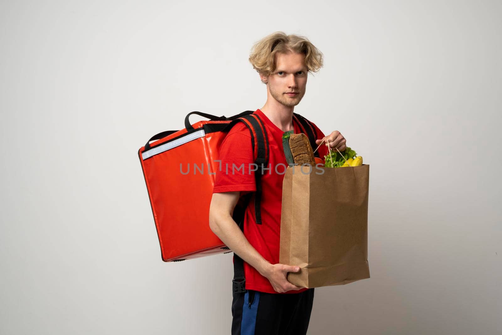 Food delivery service. Portrait of pleased delivery man in red uniform smiling while carrying paper bag with food products isolated over white background