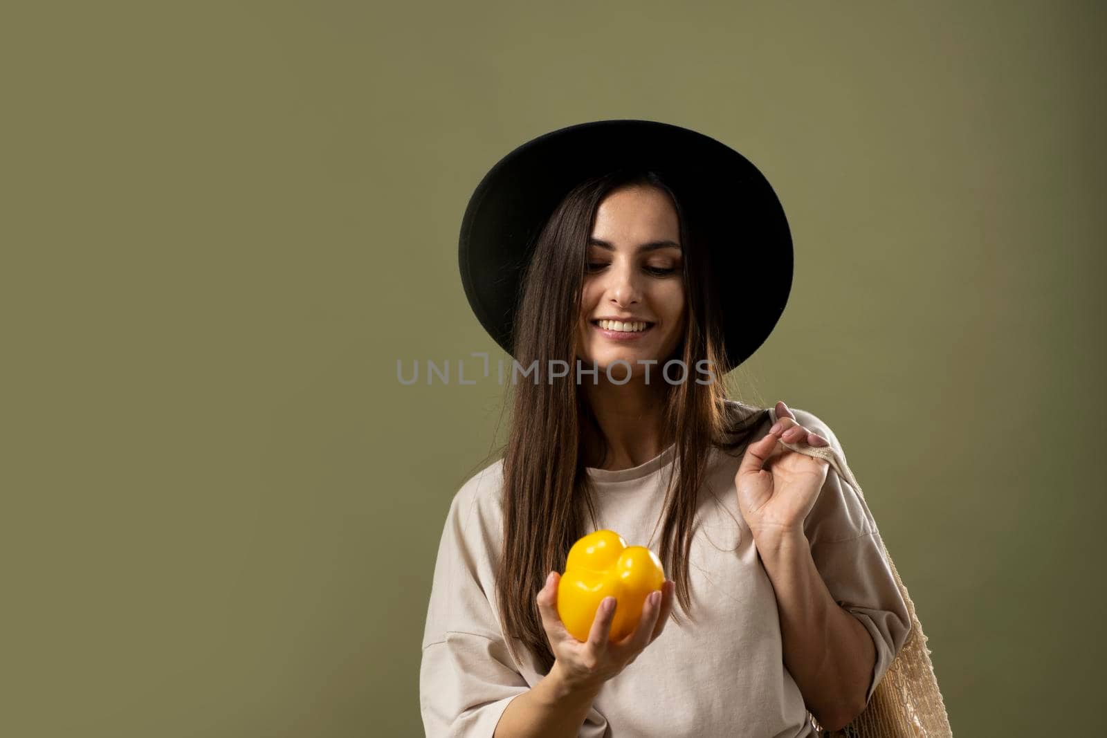 Eco friendly smiling woman in beige t-shirt and black hat holding reusable cotton eco bags full of groceries on a shoulder and yellow pepper