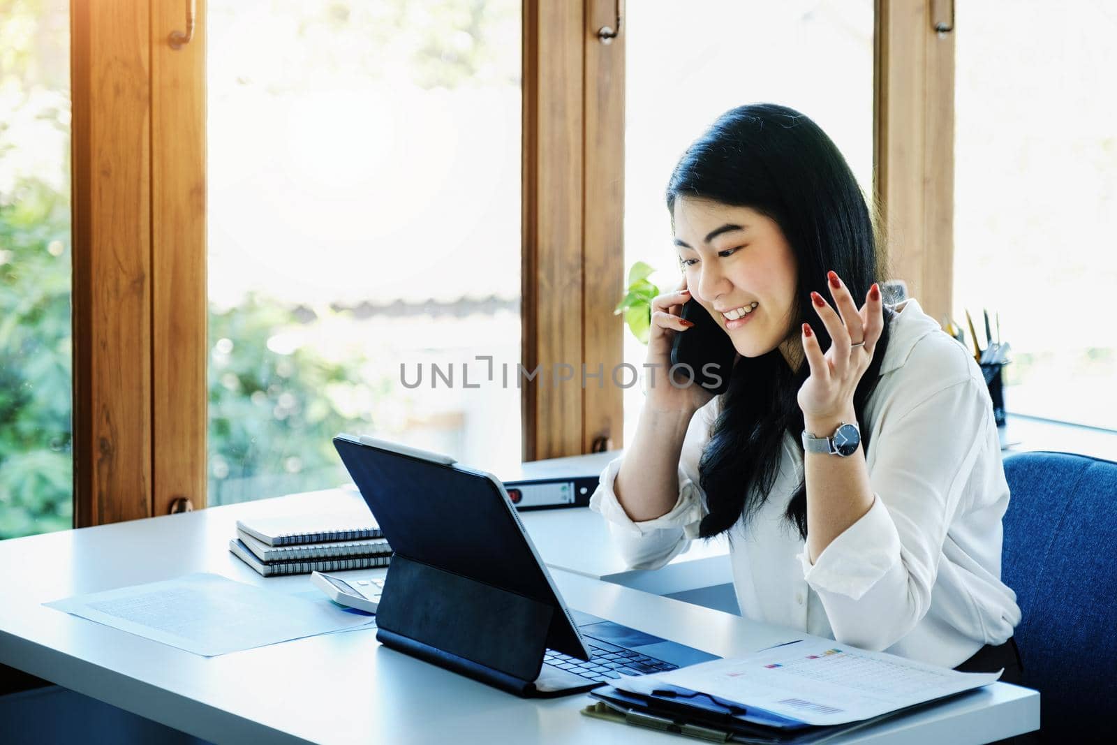 A female entrepreneur or businesswoman shows a smiling face while using smartphone and tablet computer for video conference working on a wooden table. by Manastrong