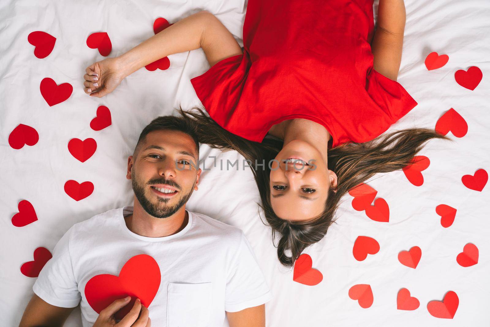 Top view of happy young couple in love with red paper hearts lying on the white bed. Love and relationship by DariaKulkova