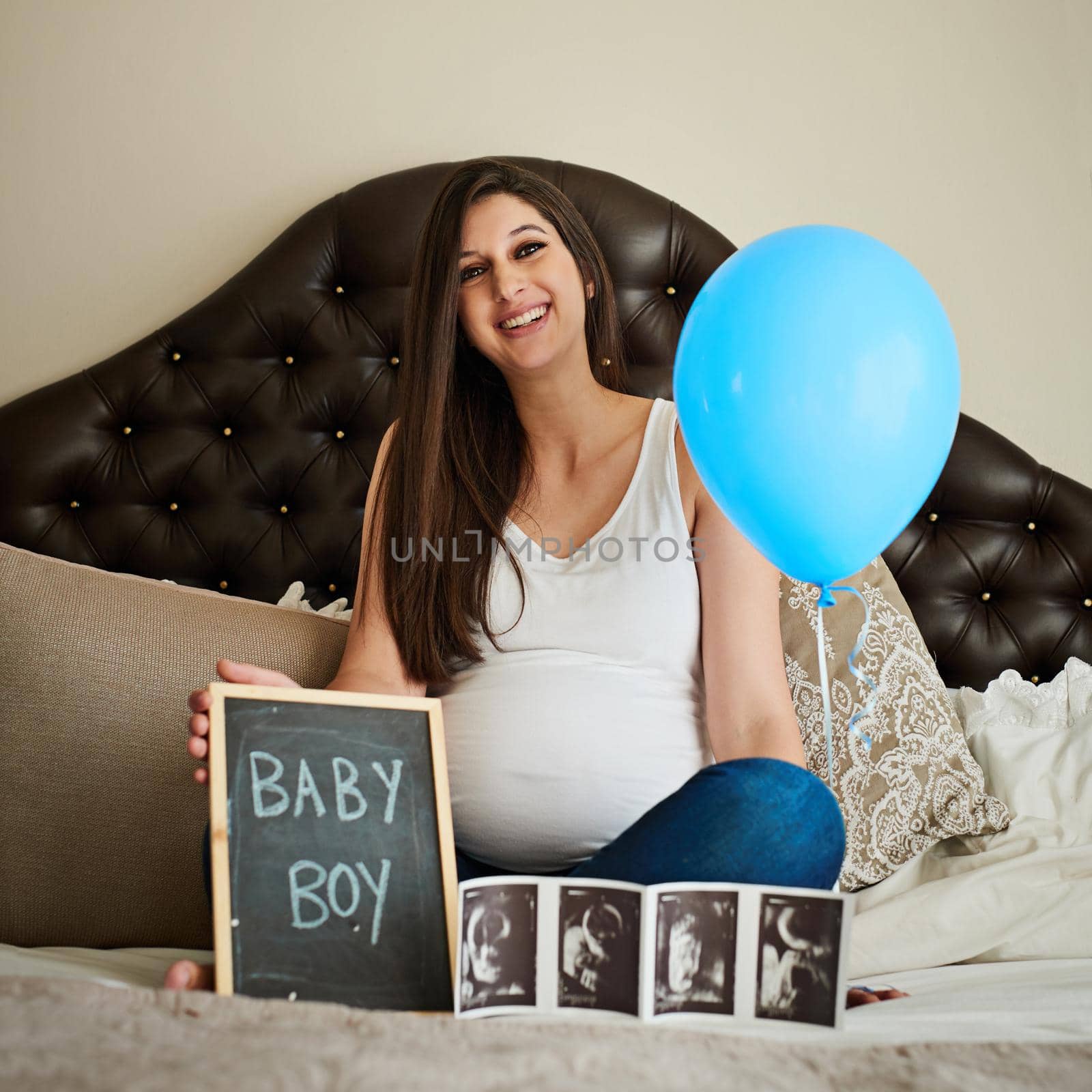 Portrait of a happy pregnant woman posing on her bed with an ultrasound picture, chalkboard and blue balloon.