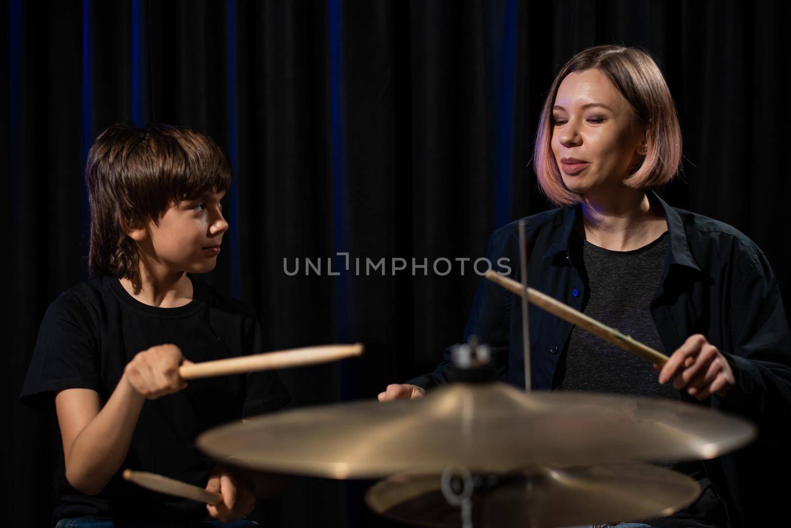 Young caucasian woman teaches a boy to play the drums in the studio on a black background. Music school student.