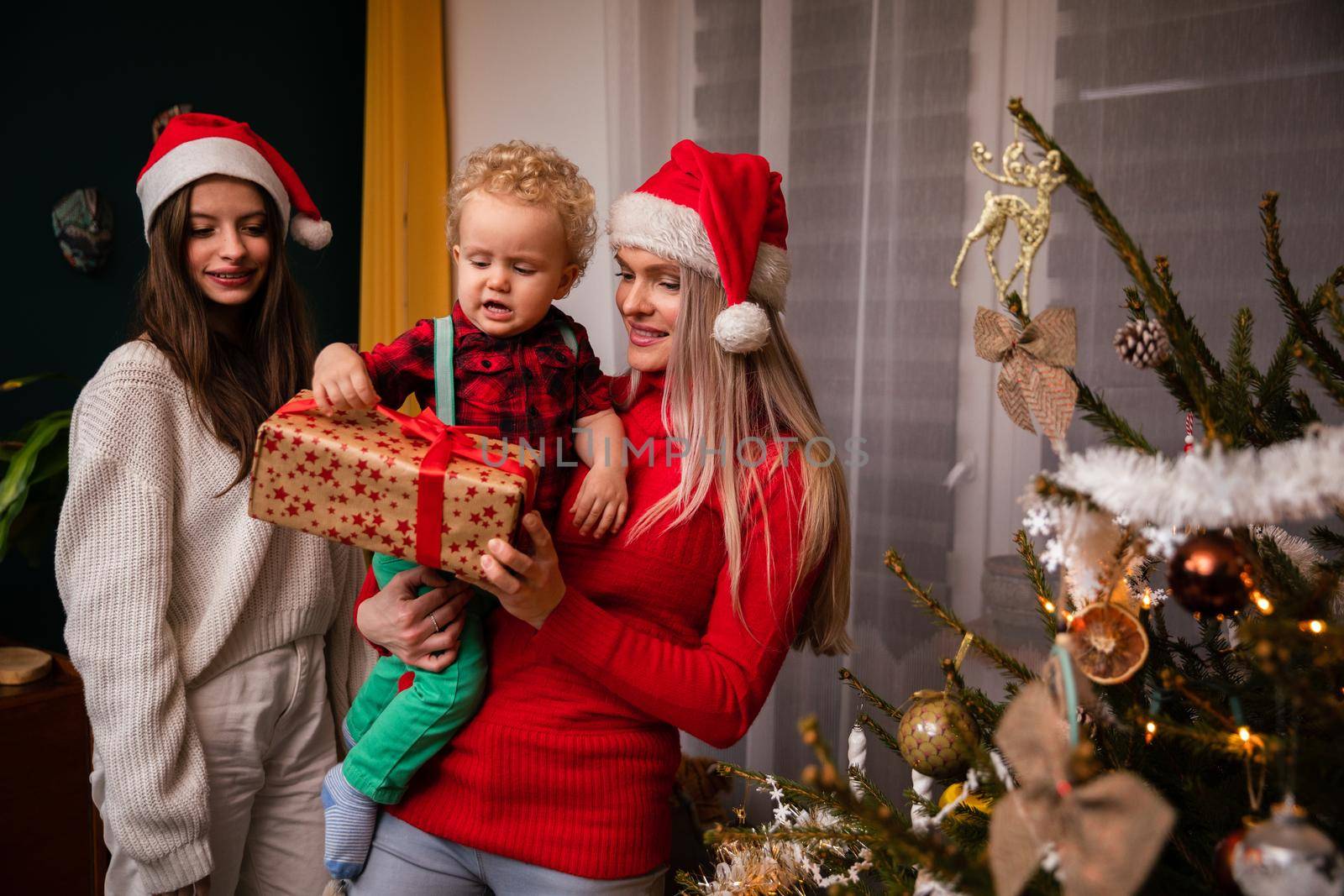 Parent with son in arms and sister give gift on Christmas Eve. Mother and daughter wearing a red Santa hat.