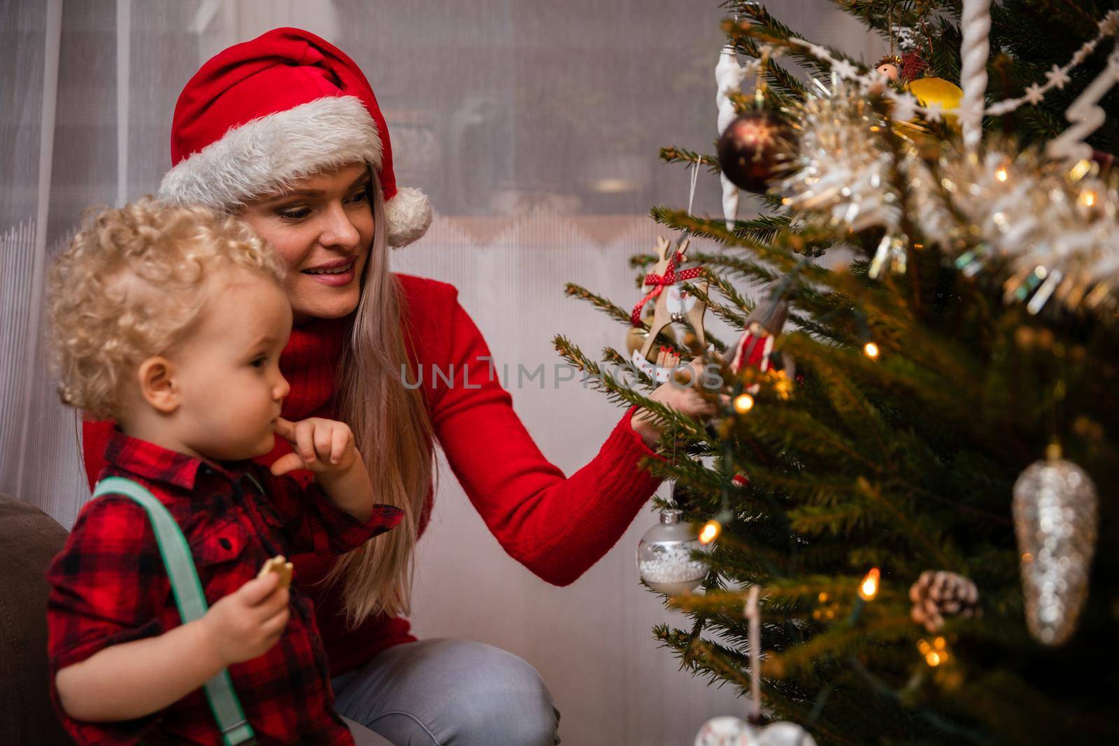 A young child holds her finger in her mouth and her mother talks about the history and traditions of Christmas. A woman wearing a red Santa hat.