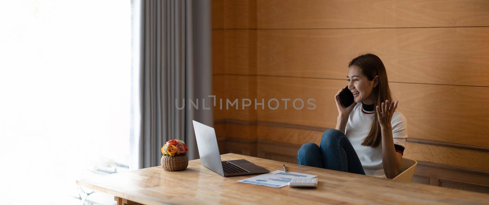 Happy young asian woman talking on the mobile phone and smiling while sitting at her working with laptop and paperwork at home.