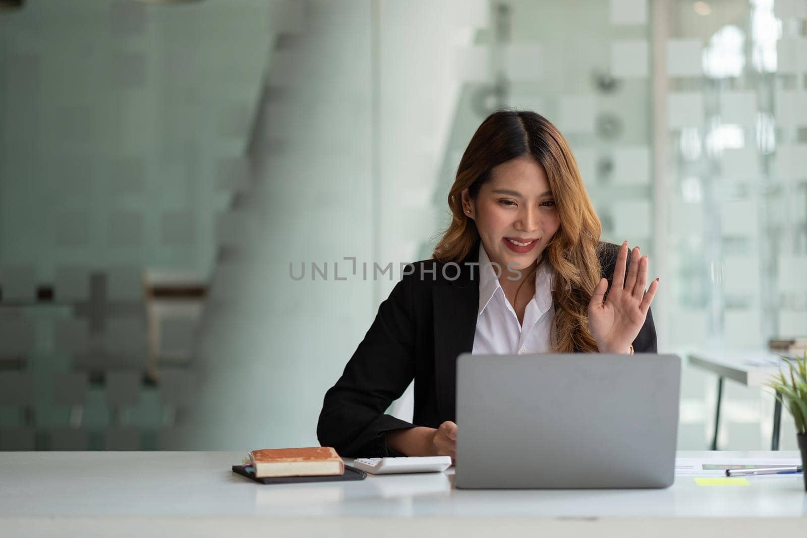 Portrait of smiling asian woman waving hello talking on video call. Successful young woman sitting white suits. Business conference via laptop.