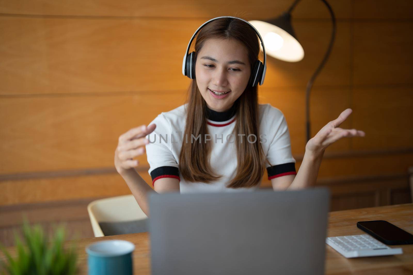 Smiling young asian woman in headset wave greet talking on webcam virtual conversation on laptop, happy female in wireless headphones speak on video call on computer, consult client online.