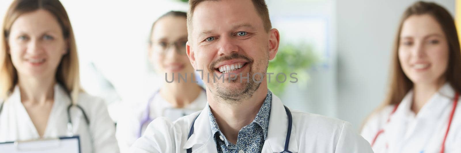 Focus on smiling male therapist offer handshake. Team of physicians in white gowns with stethoscopes posing in hospital. Medicine and healthcare concept