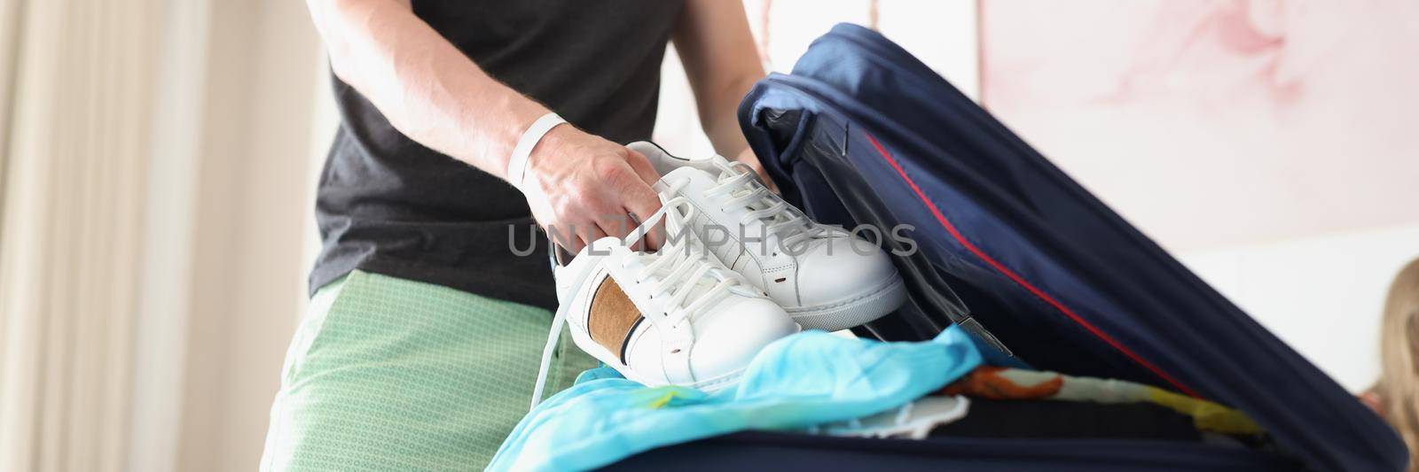 Portrait of man packing clothes into travel bag, putting pair of white sneakers into suitcase. Get ready for travel fast. Moving, traveling, business trip, relocation concept