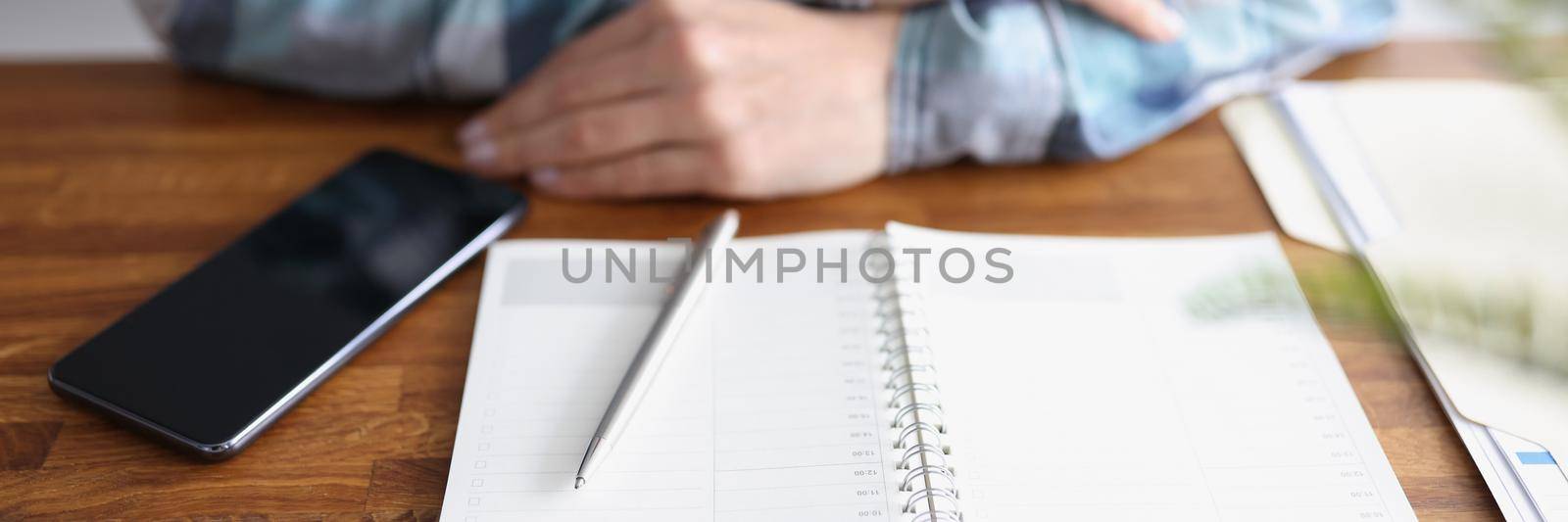 Close-up of open planner on wooden table, person with folded arms. Need to start planning week, meetings, write down thoughts and ideas. Personal diary, creative, journaling concept. Copy space