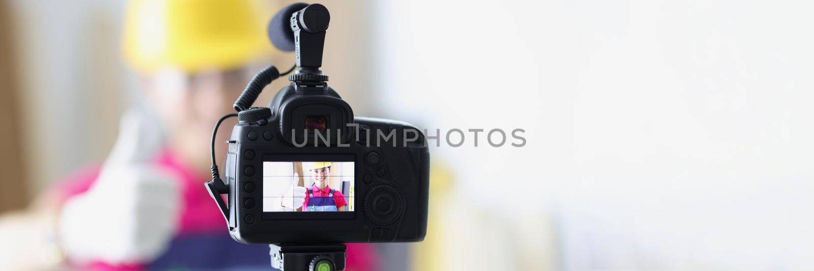 Close-up of videocamera with female repairer in helmet showing thumb up gesture to camera. Building blogging and vlogging concept. Redecoration online idea
