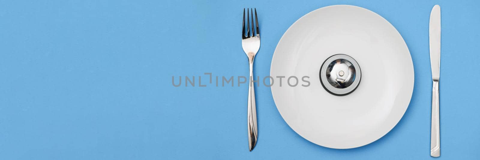 Top view of white plate with iron bell and appliances for food. Time to eat, dieting and meal-planning concept. Meals on schedule idea. Isolated on blue background