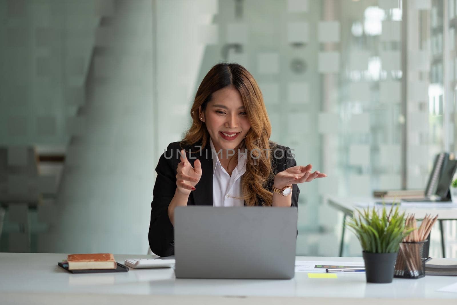 Portrait of smiling asian woman waving hello talking on video call. Successful young woman sitting white suits. Business conference via laptop.