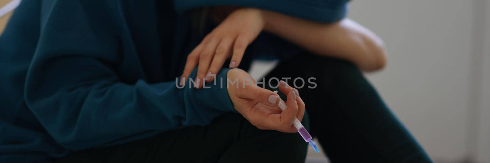 Close-up of female in hoodie holding syringe and getting high. Drug addict person stoned after narcotic injection sitting on floor. Drug habit and meth-head concept