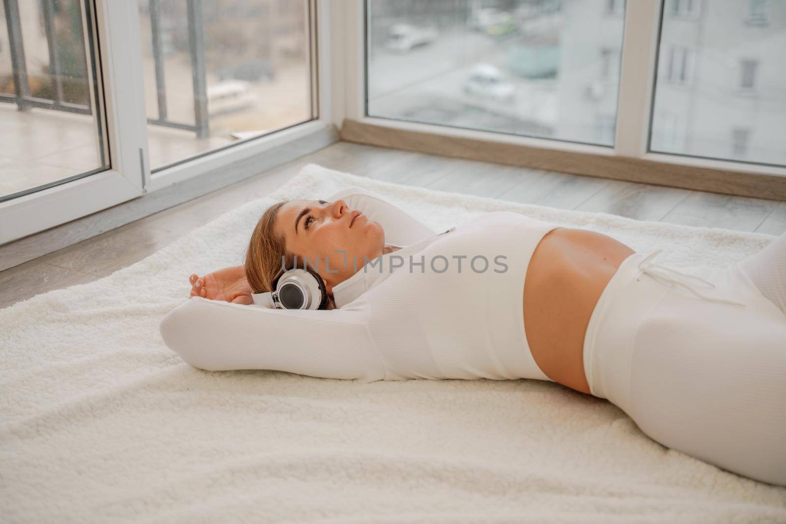 Side view portrait of relaxed woman listening to music with headphones lying on carpet at home. She is dressed in a white tracksuit. by Matiunina