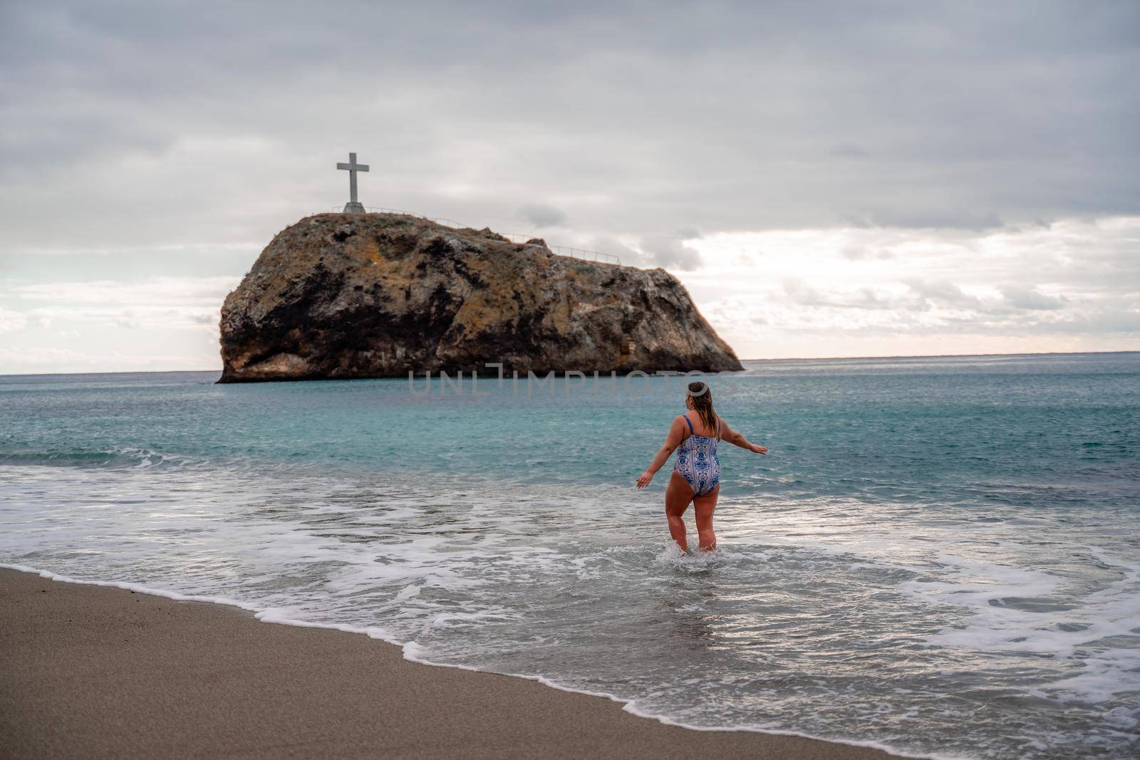 A plump woman in a bathing suit enters the water during the surf. Alone on the beach, Gray sky in the clouds, swimming in winter