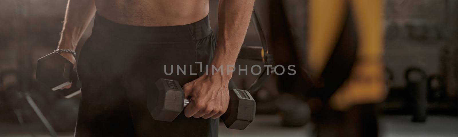 Cropped view of muscular young man holding dumbbells in hands and doing exercise to strengthen his biceps in fitness center