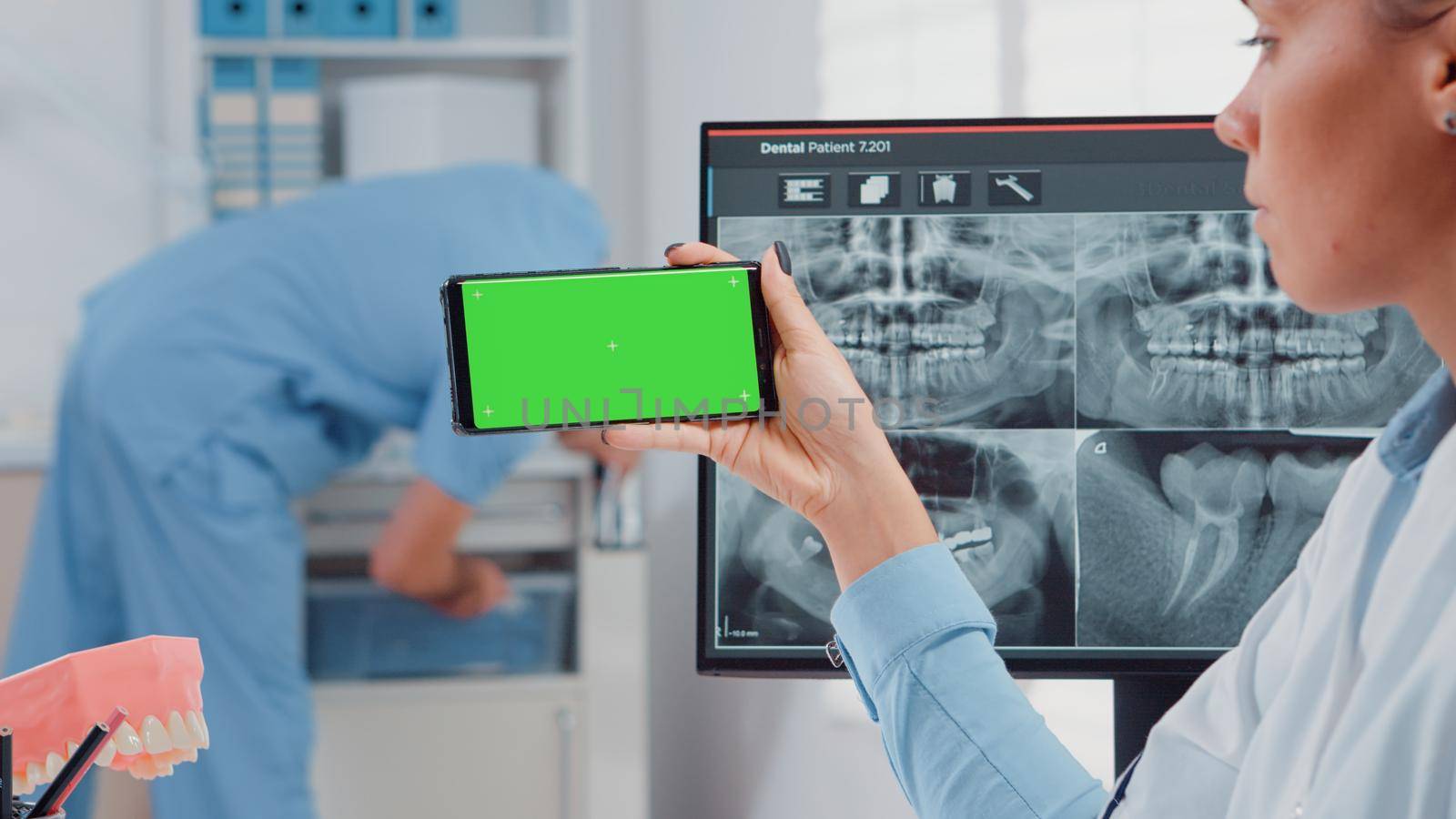 Close up of dentist analyzing smartphone with green screen and teeth scan on computer in dental cabinet. Woman holding device with chroma key for isolated mockup template for dentistry