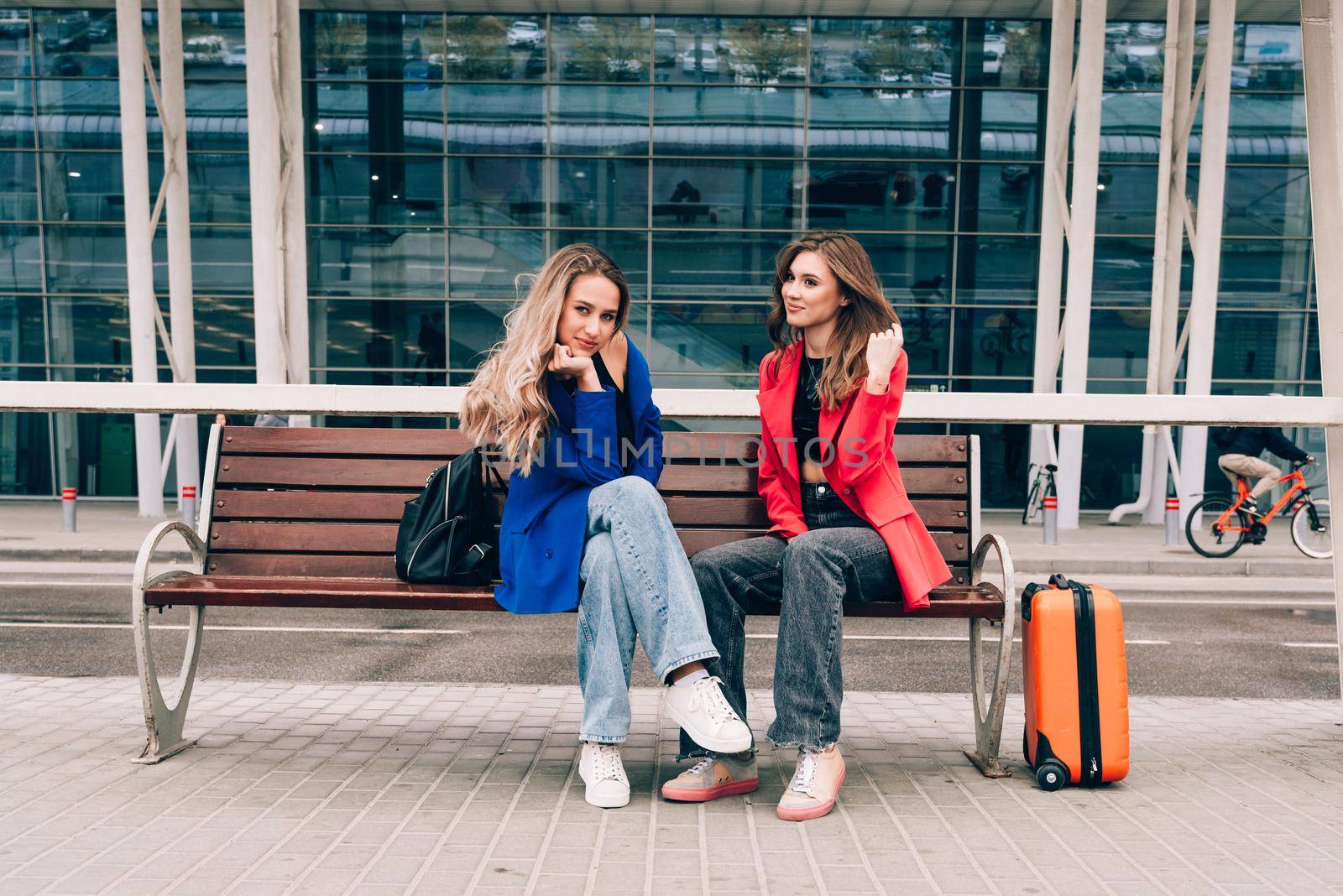 Two happy girls sitting on a bench near airport, with luggage. Air travel, summer holiday by Ashtray25