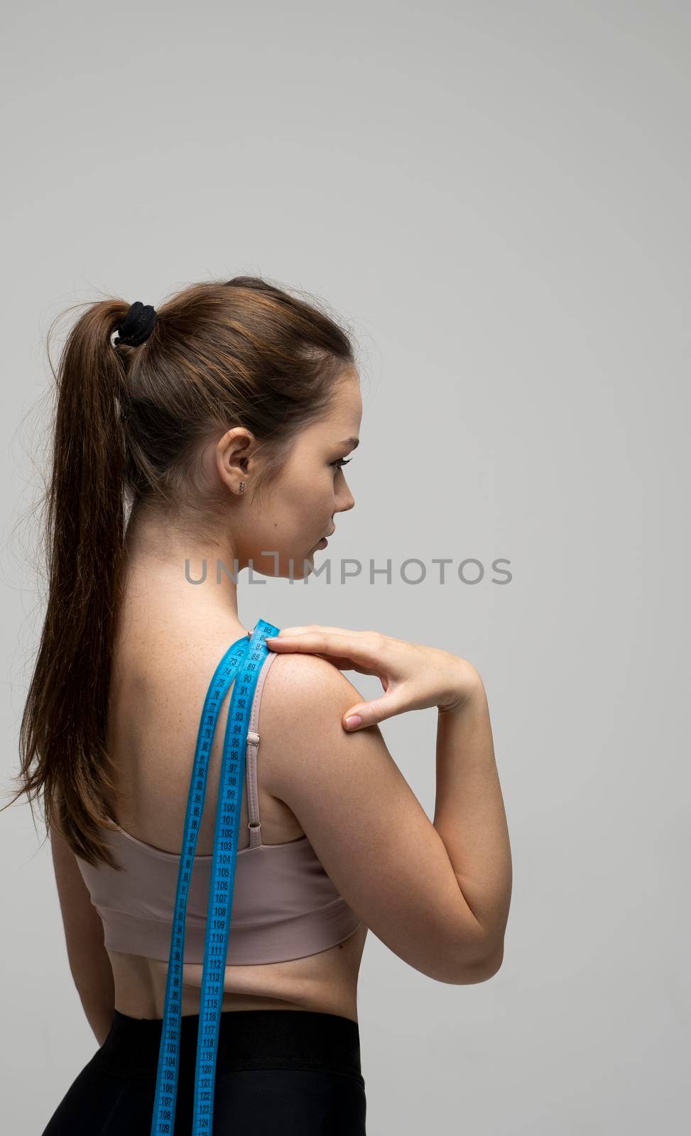 Back of young beautiful sporty brunette woman in sportswear outfit after fitness trainings with a blue measuring tape on a shoulder. Weight loss and diet concepts. by vovsht