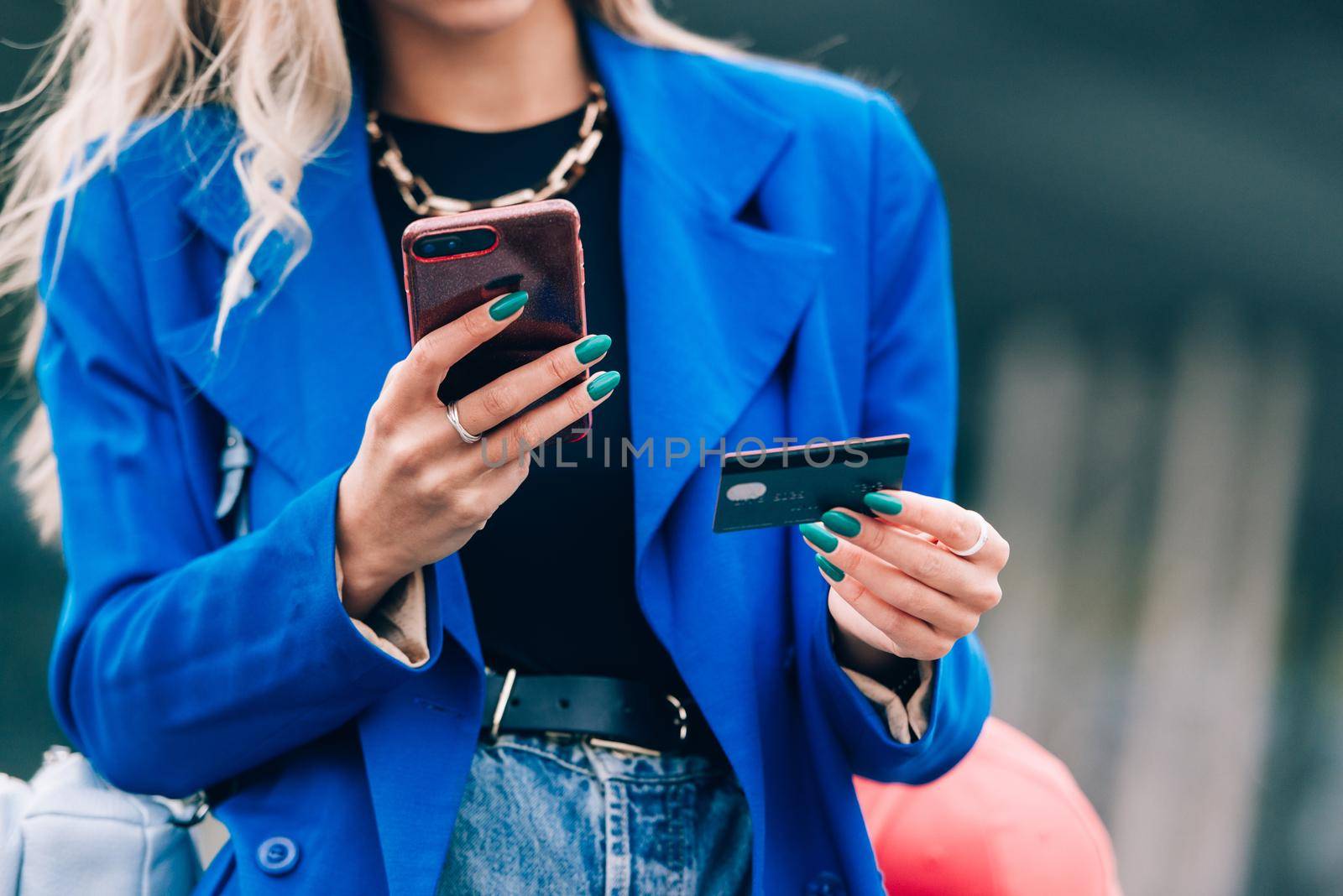 Happy fashion blonde shopper wearing a blue jacket paying on line with a credit card and a smart phone on the street.