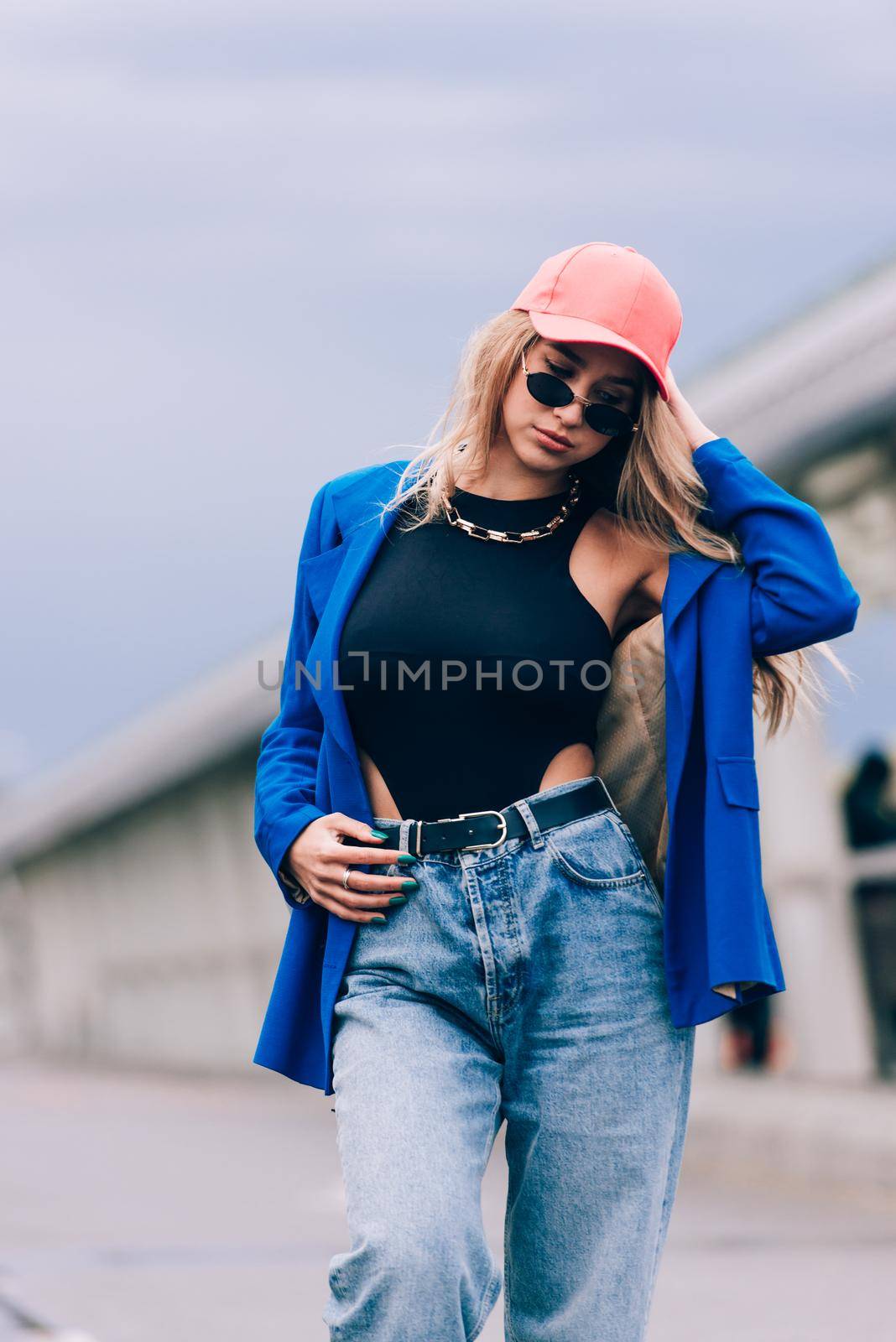 Young sexy blonde hipster woman posing on the street. Wearing blue stylish jacket, jeans and baseball hat and sunglasses. Lifestyle portrait