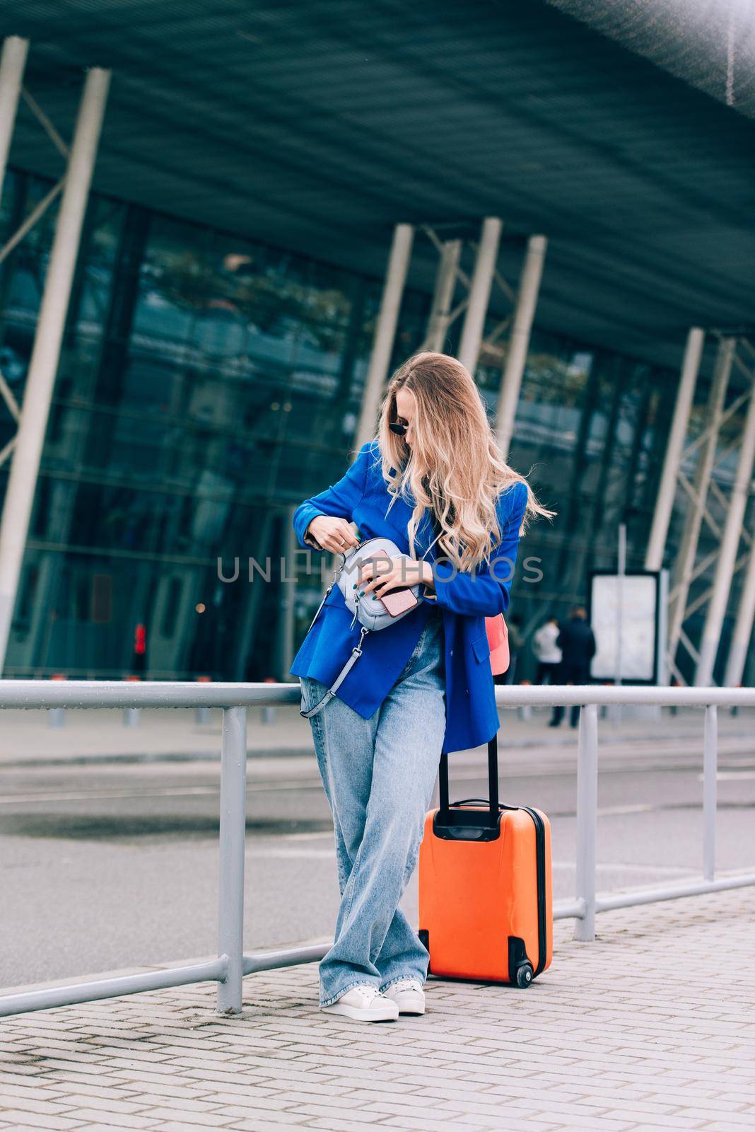 Portrait of a traveler woman standing with an orange suitcase near an airport. Young fashionable woman in a blue jeans and jacket, black shirt and white sneakers by Ashtray25